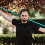 Adult man in black shirt exercises with a resistance band outside.